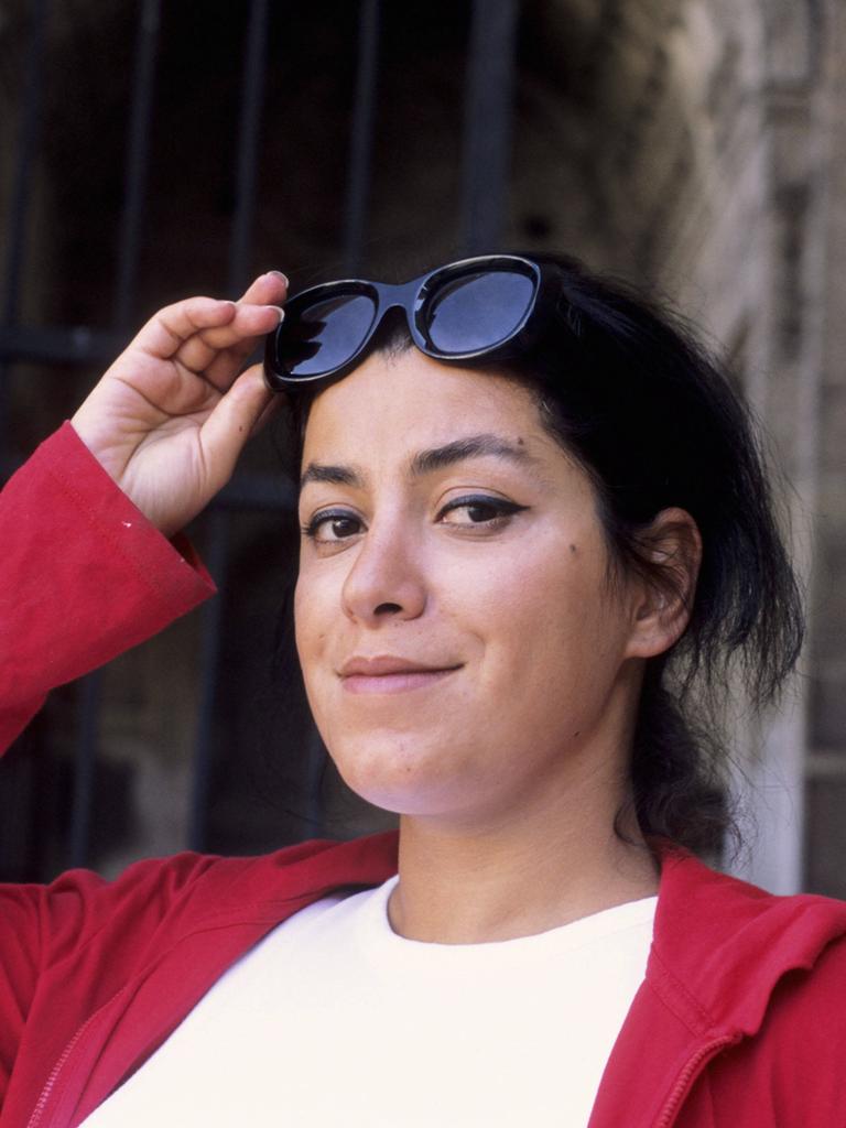 Die Künstlerin Marjane Satrapi im Porträt. Sie schaut verschmitzt in die Kamera, hält ihre Sonnenbrille fest und trägt eine rote Jacke