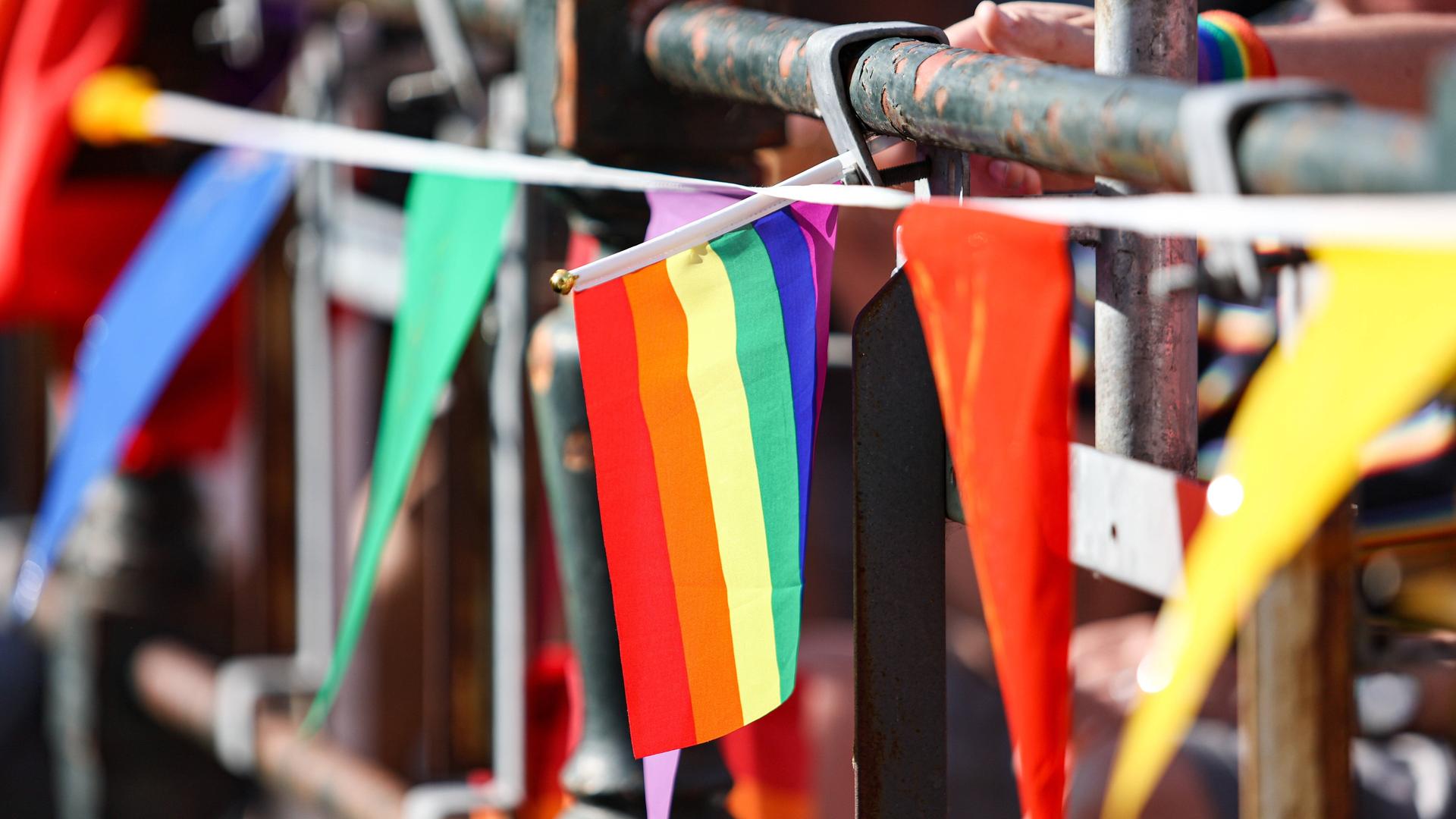 An einem Brückengeländer sind verschiedene Bänder und Fahnen in bunten Farben befestigt. Im Bildzentrum: Die Regenbogenfahne der LGBTQ-Community. 