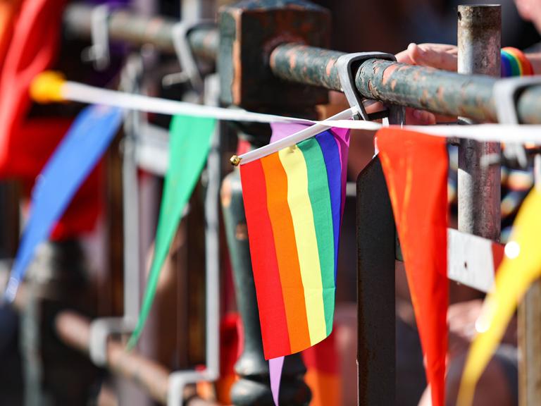 An einem Brückengeländer sind verschiedene Bänder und Fahnen in bunten Farben befestigt. Im Bildzentrum: Die Regenbogenfahne der LGBTQ-Community. 
