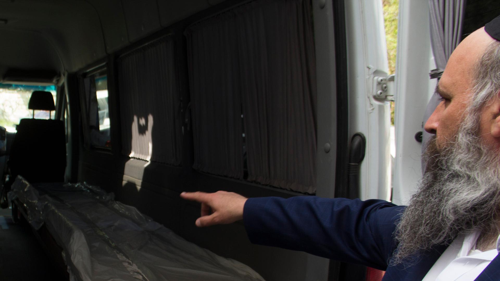 Rabbi Mayer Stambler. Ein älterer Herr mit Vollbart und Kippa zeigt auf zwei Liegen im Innenraum eines Transporters. 