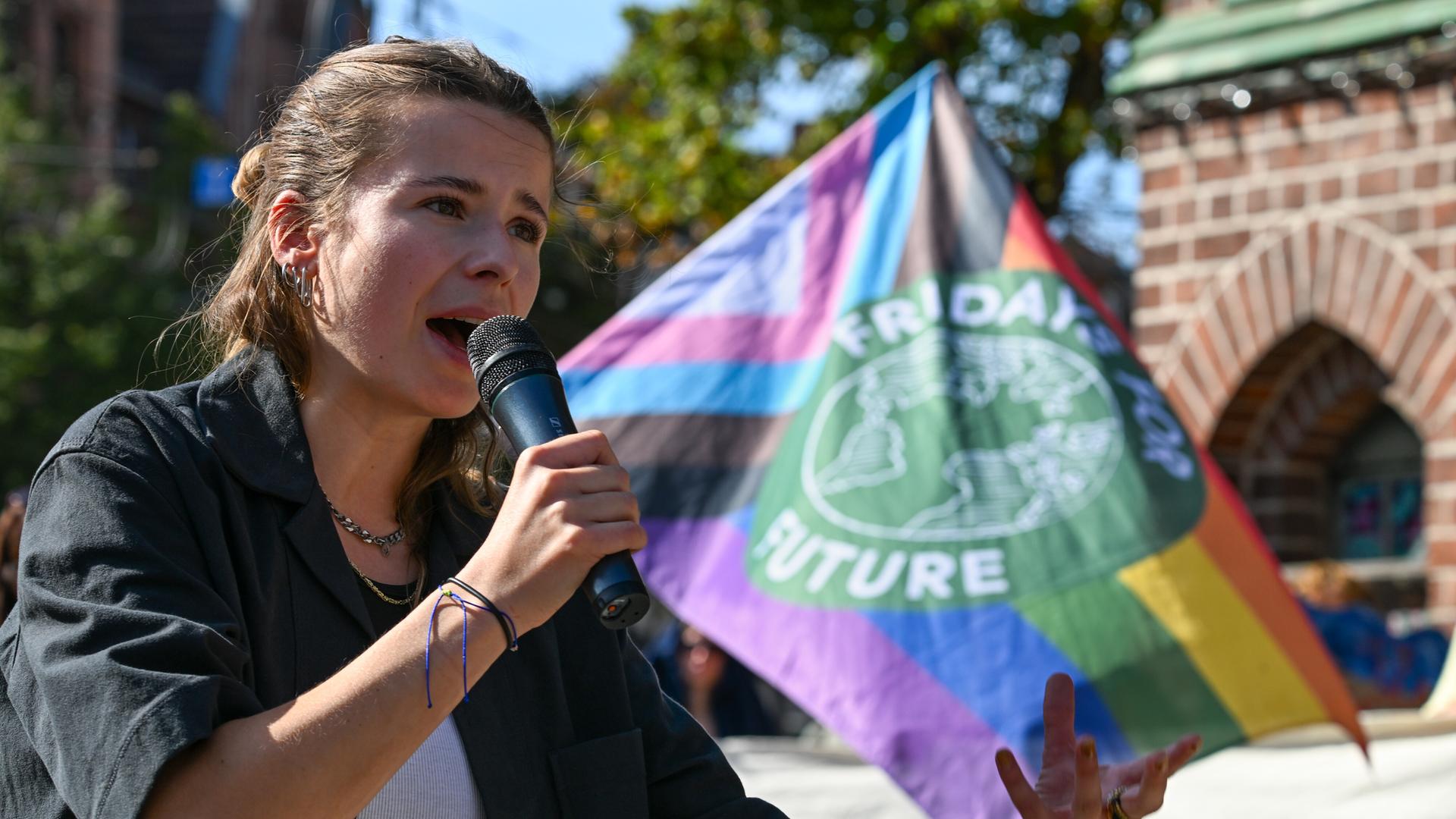 Luisa Neubauer von Fridays for Future spricht während einer Demonstration vor dem Babelsberger Rathaus. Sie trägt eine dunkle Bluse aus grobem Stoff und spricht in ein Mikrofon. Rechts im Bild die Flagge der Klimaschutzbewegung. Luisa Neubauer von Fridays for Future spricht während einer Demonstration vor dem Babelsberger Rathaus. Sie trägt eine dunkle Bluse aus grobem Stoff und spricht in ein Mikrofon. Rechts im Bild die Flagge der Klimaschutzbewegung.