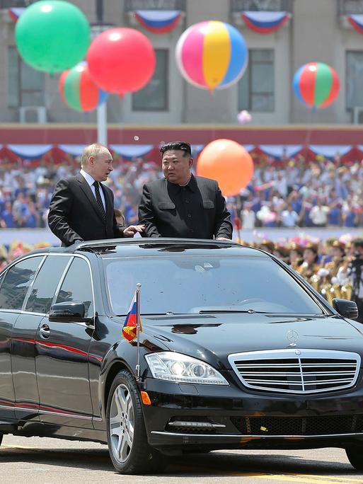 Russlands Präsident Putin und Nordkoreas Machthaber Kim Jong Un stehen im geöffneten Dachfenster eines schwarzen Autos, das durch eine Willkommensparade in Pjöngjang fährt. Im Hintergrund sind bunte Ballons und viele Menschen zu sehen. 