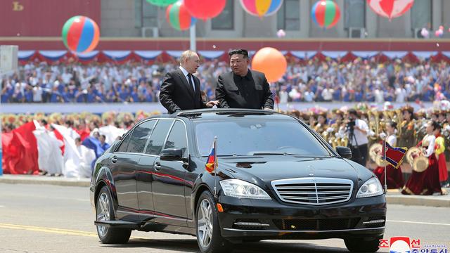 Russlands Präsident Putin und Nordkoreas Machthaber Kim Jong Un stehen im geöffneten Dachfenster eines schwarzen Autos, das durch eine Willkommensparade in Pjöngjang fährt. Im Hintergrund sind bunte Ballons und viele Menschen zu sehen. 