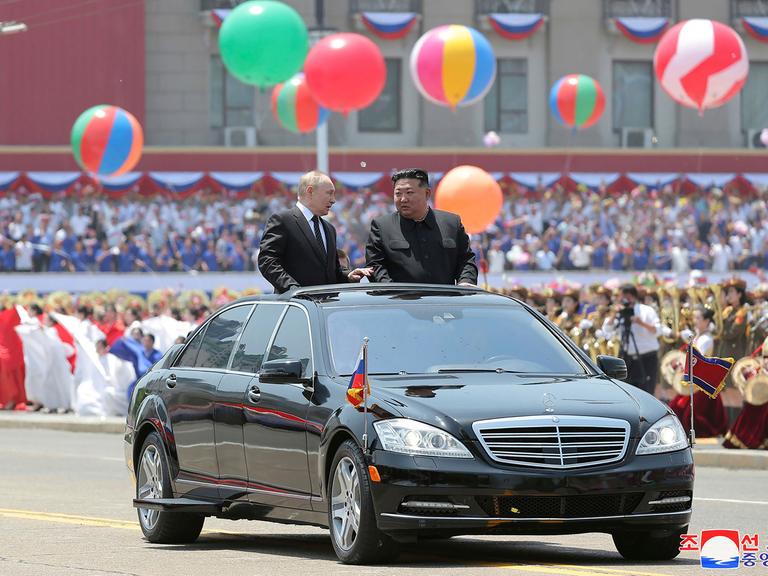 Russlands Präsident Putin und Nordkoreas Machthaber Kim Jong Un stehen im geöffneten Dachfenster eines schwarzen Autos, das durch eine Willkommensparade in Pjöngjang fährt. Im Hintergrund sind bunte Ballons und viele Menschen zu sehen. 