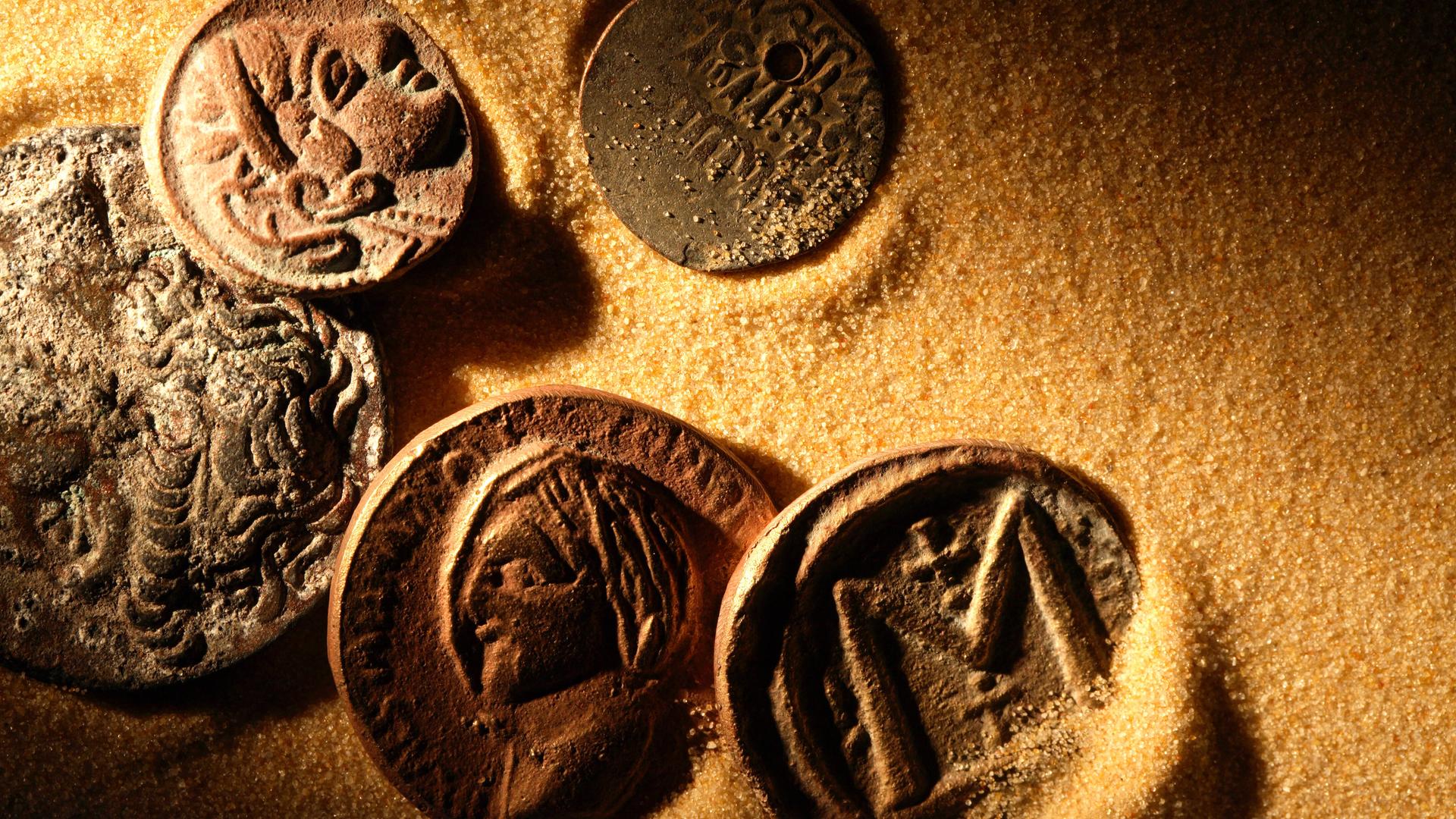 Das Bild zeigt mehrere antik aussehende Münzen, die in gelbem Sand liegen (Symbolbild)