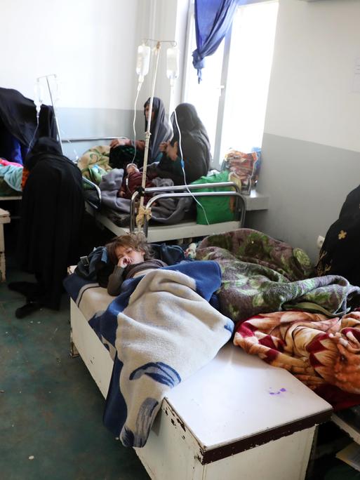 Kinder in einem Krankenhaus in Kabul. Sie liegen unter ärmlichen Decken, mehrere kleine Kinder in einem Bett, mit verhüllten Frauen die daneben sitzen.