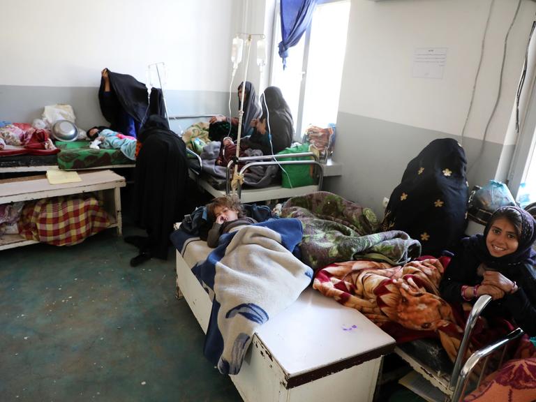 Kinder in einem Krankenhaus in Kabul. Sie liegen unter ärmlichen Decken, mehrere kleine Kinder in einem Bett, mit verhüllten Frauen die daneben sitzen.