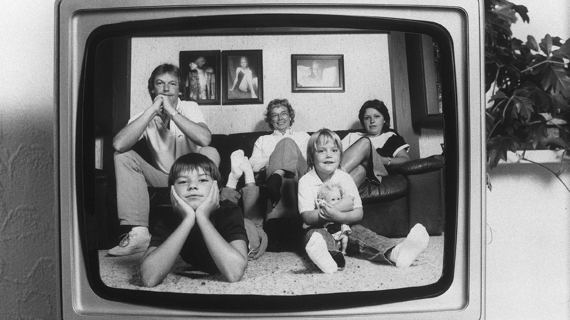 Ein Schwarz-Weiß-Foto aus den 80er-Jahren zeigt eine typische Familie auf einem Sofa, die sich in einem damals üblichen TV-Apparat spiegelt.