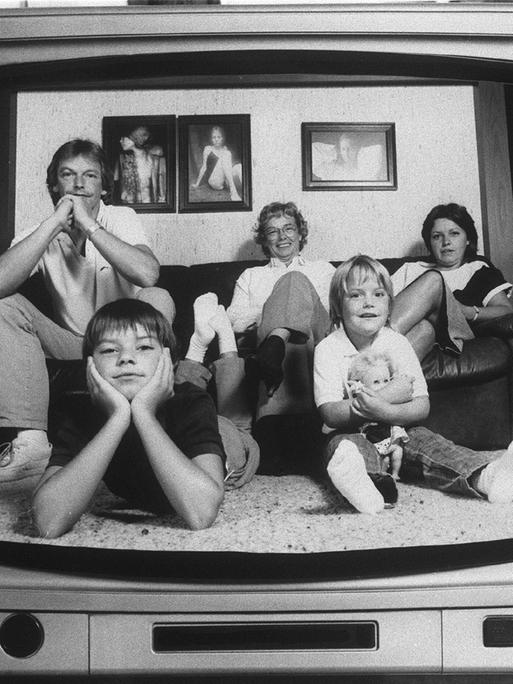 Ein Schwarz-Weiß-Foto aus den 80er-Jahren zeigt eine typische Familie auf einem Sofa, die sich in einem damals üblichen TV-Apparat spiegelt.