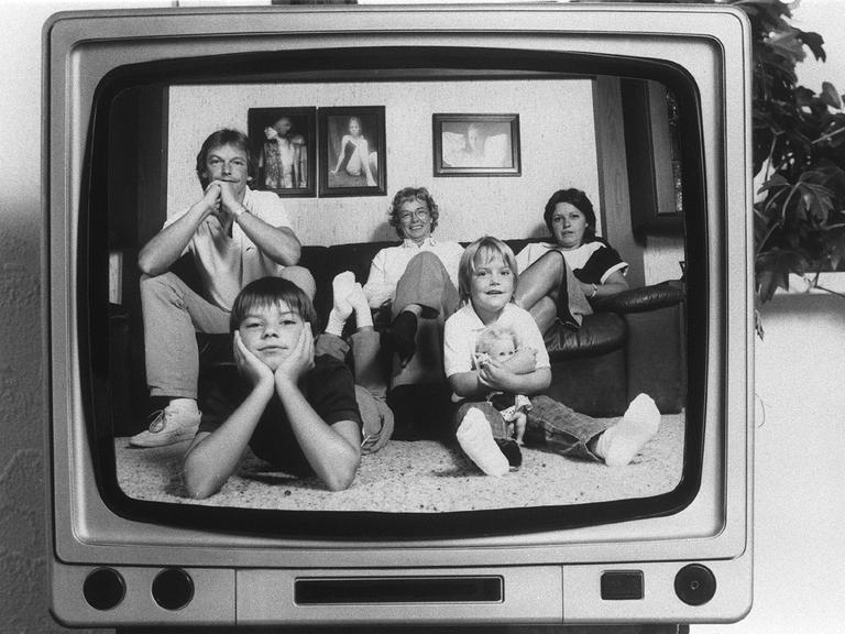 Ein Schwarz-Weiß-Foto aus den 80er-Jahren zeigt eine typische Familie auf einem Sofa, die sich in einem damals üblichen TV-Apparat spiegelt.