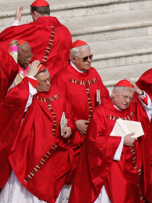 Rot gewandete Kardinäle halten auf einem Platz ihre Kappen gegen den Wind fest.