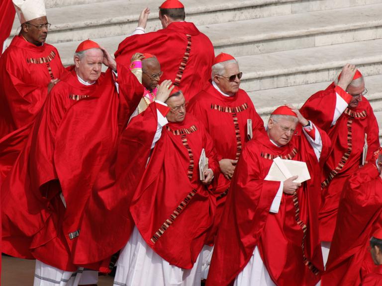 Rot gewandete Kardinäle halten auf einem Platz ihre Kappen gegen den Wind fest.