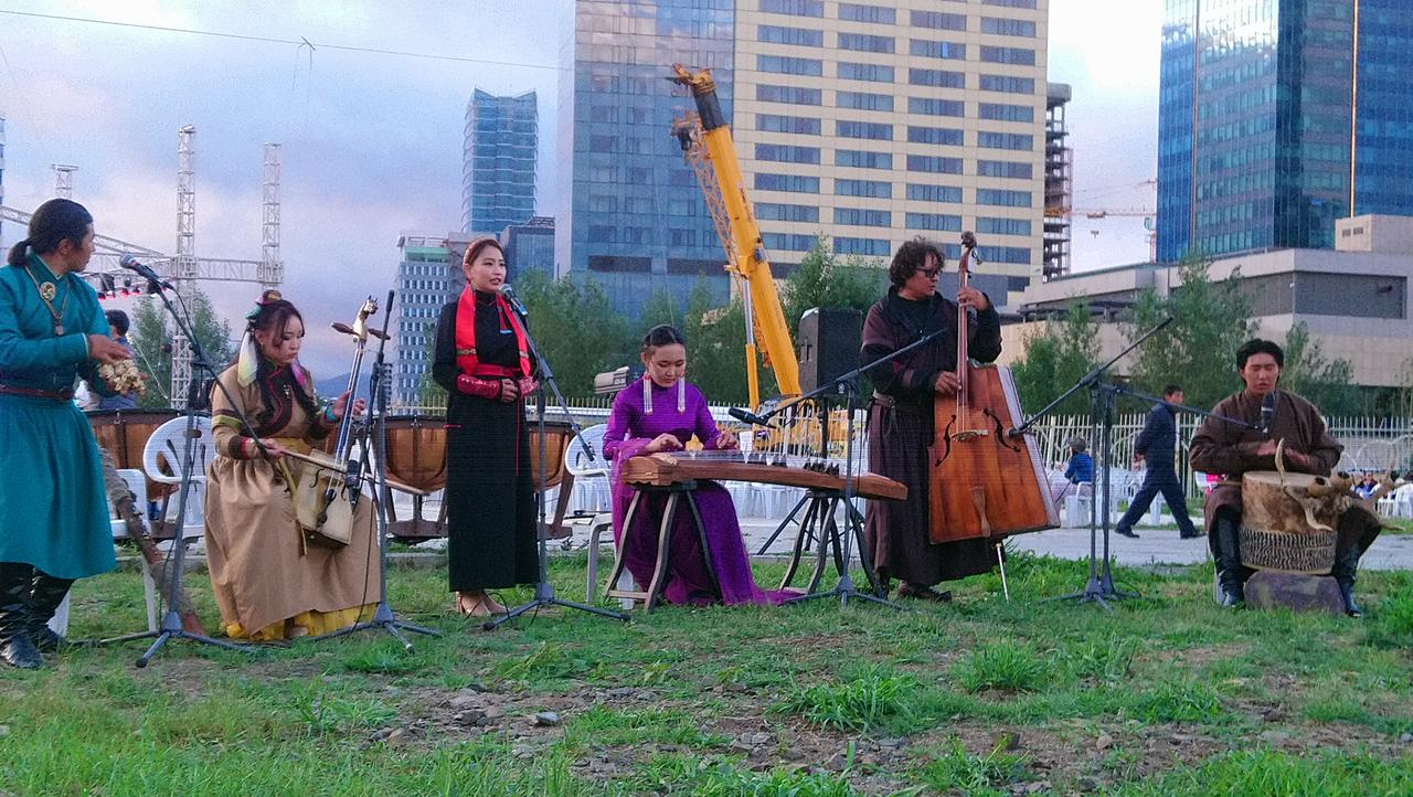 Musiker mit verschiedenen Instrumenten spielen auf einer Wiese, die vor der Hochhäusern und einem Kran liegt.