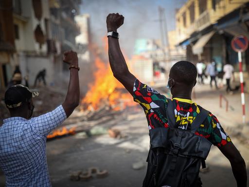 Demonstranten verbarrikadieren in Dakar eine Straße, indem sie Holz, Reifen und Eisenplatten verbrennen, um gegen die Verschiebung der Präsidentschaftswahlen im Senegal zu protestieren. Im Vordergrund sind zwei Männer von der Rückenansicht mit erhobenen Armen zu sehen. Weiter hinten lodern Flammen. 