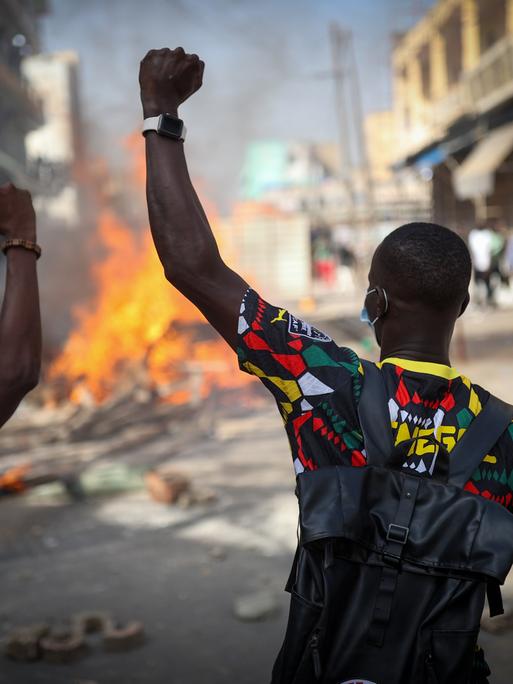 Demonstranten verbarrikadieren in Dakar eine Straße, indem sie Holz, Reifen und Eisenplatten verbrennen, um gegen die Verschiebung der Präsidentschaftswahlen im Senegal zu protestieren. Im Vordergrund sind zwei Männer von der Rückenansicht mit erhobenen Armen zu sehen. Weiter hinten lodern Flammen. 