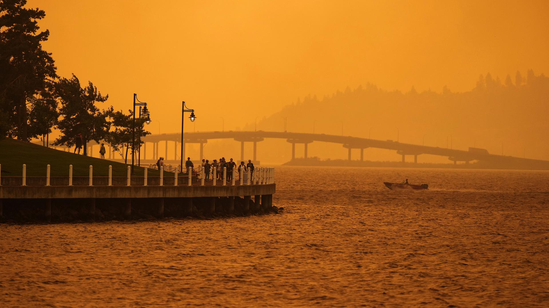 Eine Person fährt in einem Boot an Menschen vorbei, die auf einer Promenade spazieren gehen, während sich der Rauch eines Waldbrandes am Okanagan Lake in Kelowna, British Columbia, ausgebreitet hat.