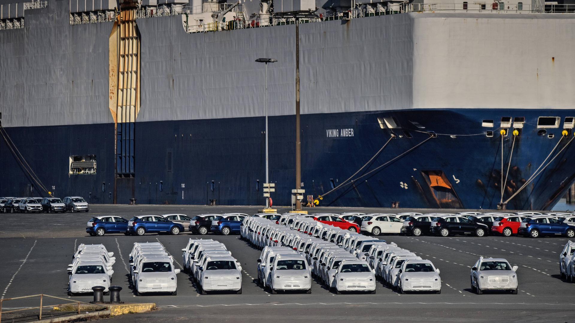 Auf dem Foto ist ein großes Schiff zu sehen. Vor dem Schiff stehen Autos. Sie haben Schutzhüllen und warten auf die Verladung auf das Schiff.