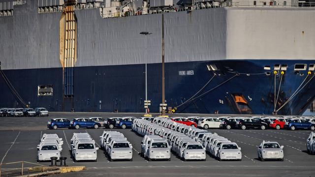Auf dem Foto ist ein großes Schiff zu sehen. Vor dem Schiff stehen Autos. Sie haben Schutzhüllen und warten auf die Verladung auf das Schiff.