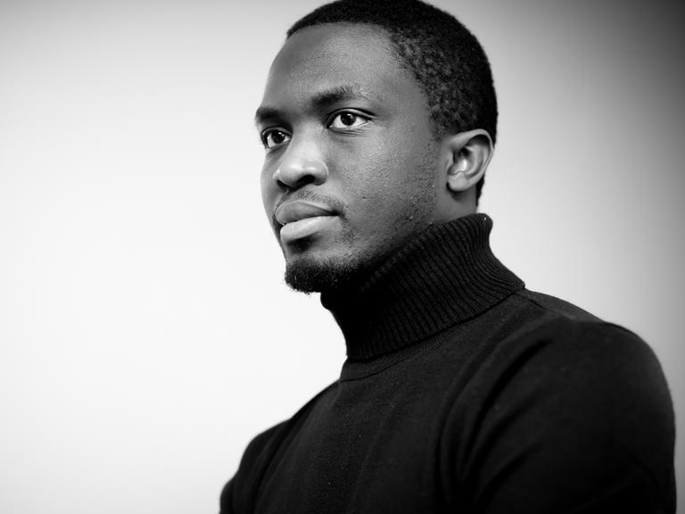 Der senegalesische Autor Mohamed Mbougar Sarr. Es handelt sich um ein Schwarzweiß-Foto, der Autor trägt einen dunklen Rollkragenpullover.