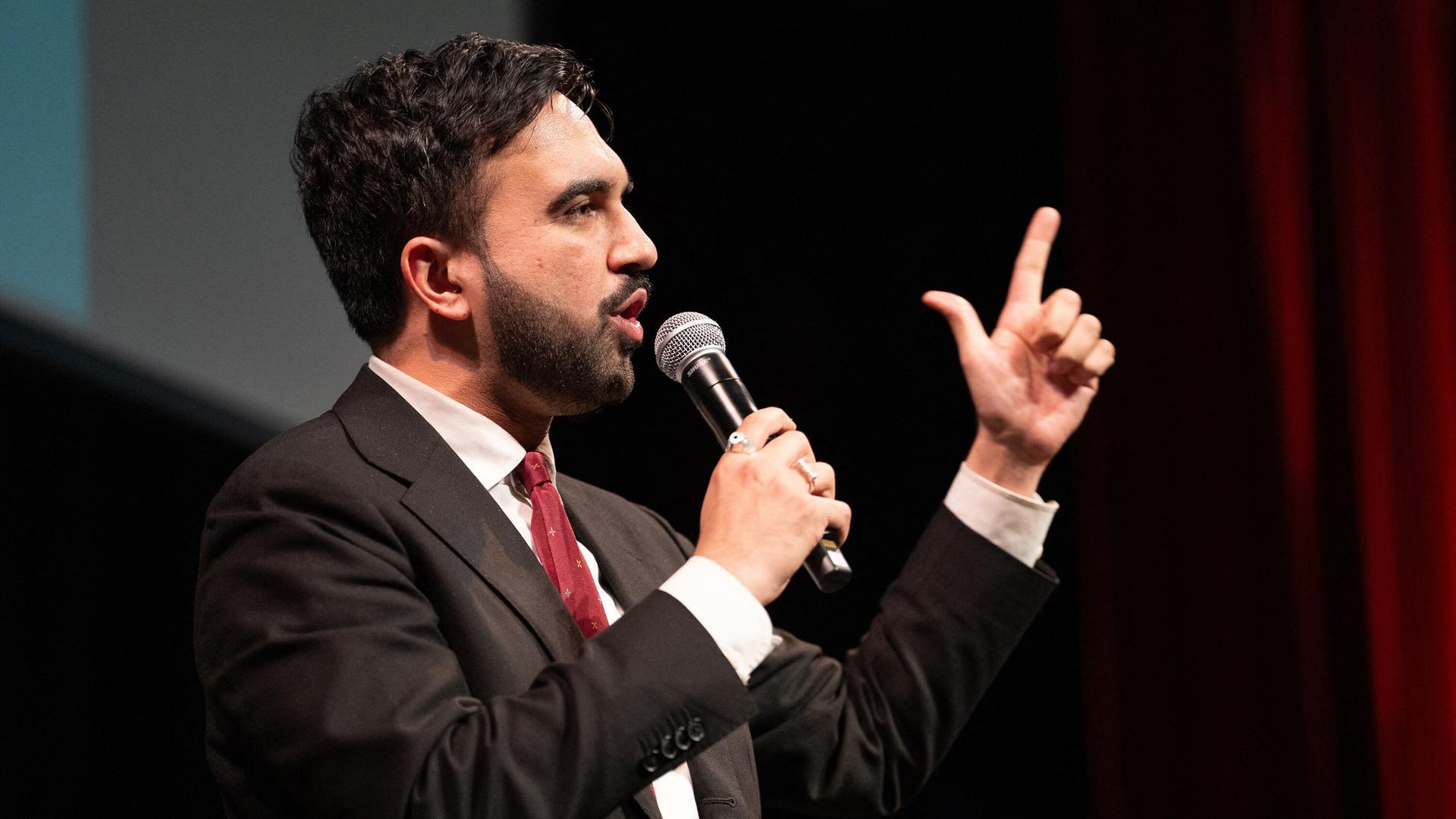 Zohran Mandani hält mit der rechten Hand ein Mikrofon, mit der linken gestikuliert er.