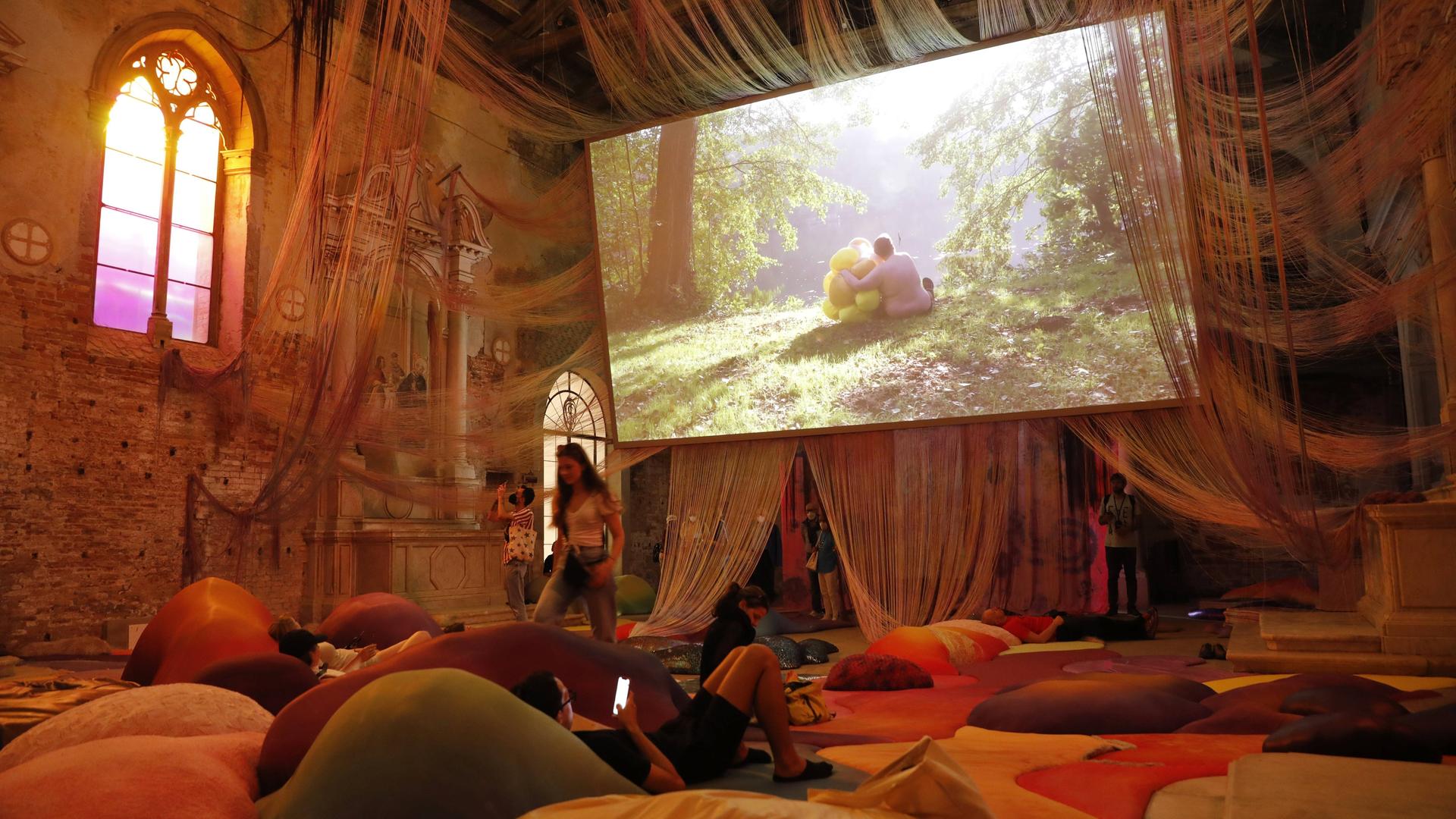 Ein mit farbigen Kissen ausgelegter Raum mit hohen Decken, von denen Schnüre hängen. Außerdem: Ein großer Bildschirm, auf dem eine Person in einer Waldlandschaft zu sehen ist.
