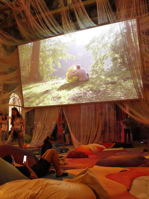 Ein mit farbigen Kissen ausgelegter Raum mit hohen Decken, von denen Schnüre hängen. Außerdem: Ein großer Bildschirm, auf dem eine Person in einer Waldlandschaft zu sehen ist.