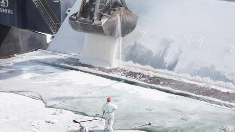 Auf einem Frachtschiff wird mit einem großen Bagger Düngemittel verladen. Im Vordergrund ist eine Person in einem weißen Schutzanzug und mit Helm zu sehen.