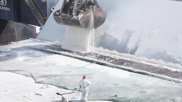 Auf einem Frachtschiff wird mit einem großen Bagger Düngemittel verladen. Im Vordergrund ist eine Person in einem weißen Schutzanzug und mit Helm zu sehen.