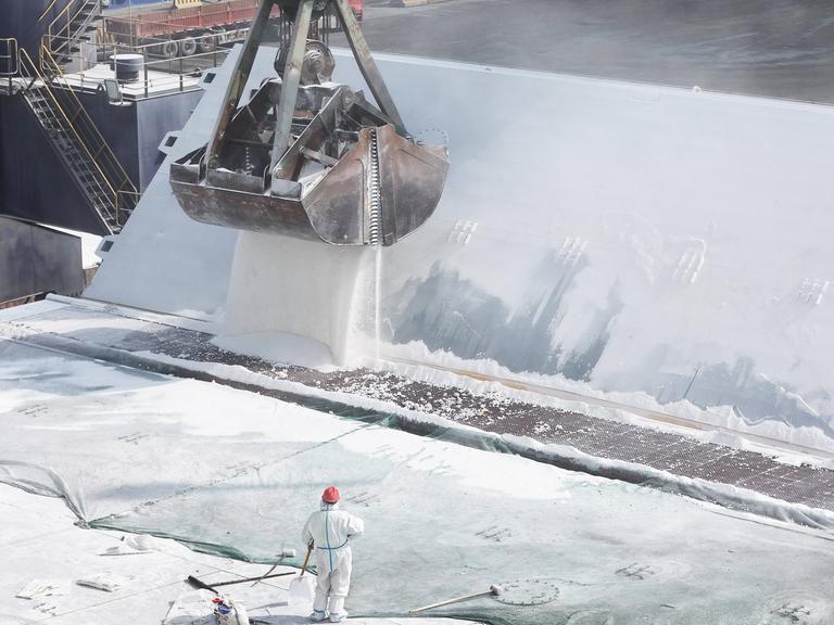 Auf einem Frachtschiff wird mit einem großen Bagger Düngemittel verladen. Im Vordergrund ist eine Person in einem weißen Schutzanzug und mit Helm zu sehen.