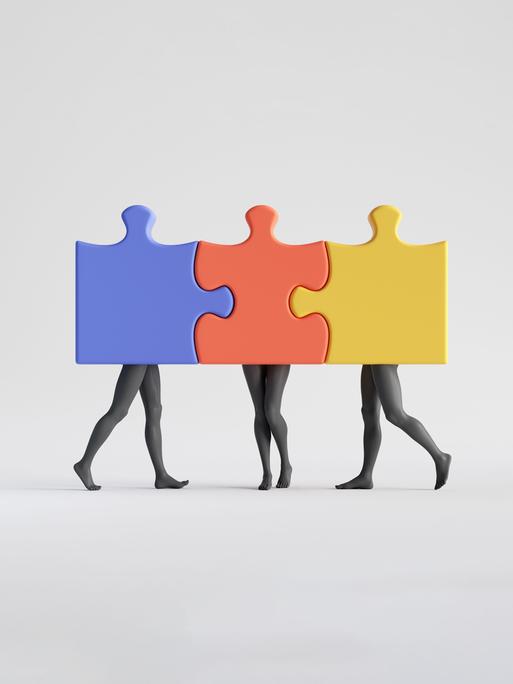 Schwarz-Weiß-Aufnahme dreier Personen, deren Beine zu sehen sind und deren Oberkörper hinter farbigen, ineinandergesteckten Puzzleteilen verborgen sind.