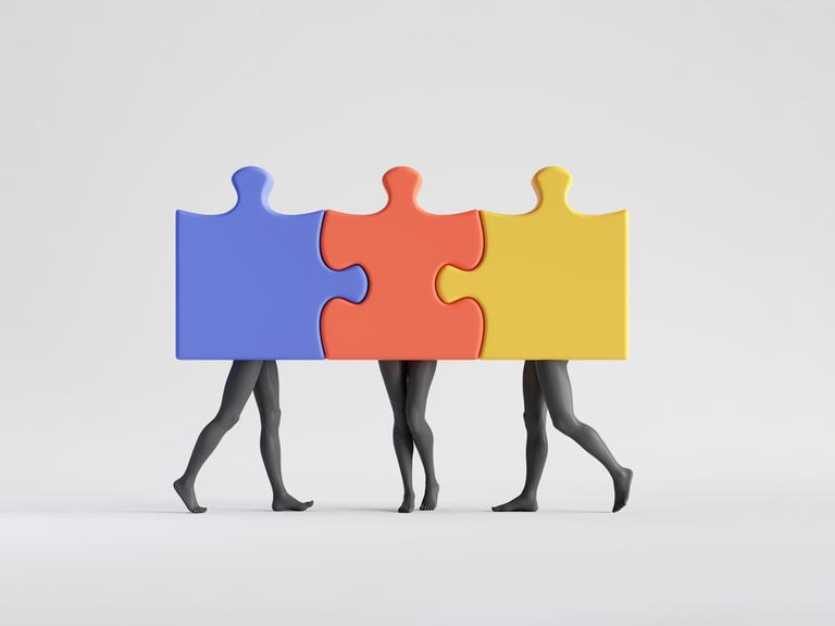 Schwarz-Weiß-Aufnahme dreier Personen, deren Beine zu sehen sind und deren Oberkörper hinter farbigen, ineinandergesteckten Puzzleteilen verborgen sind.