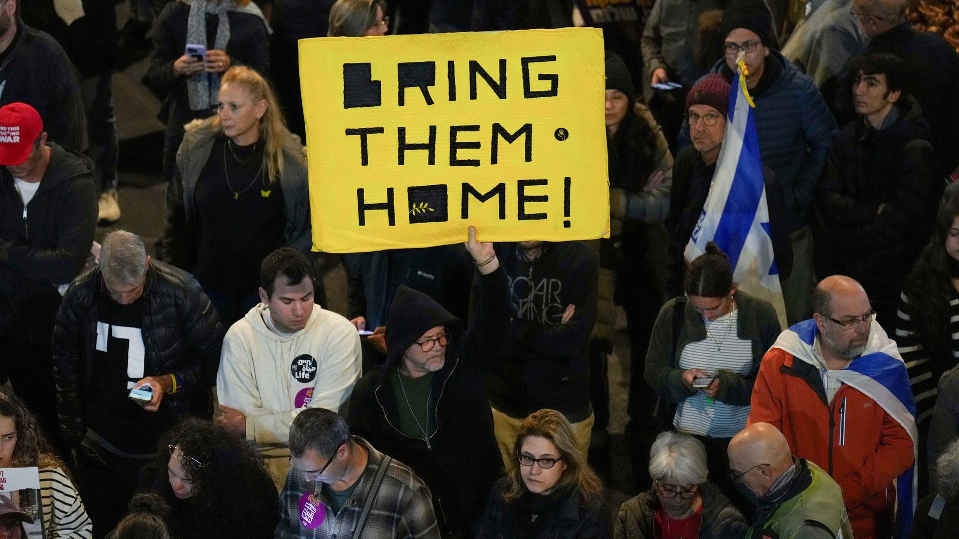 Es ist dunkel. Das Bild zeigt eine Gruppe Demonstranten. Auf einem gelben Pappschild steht "Bring them home". 