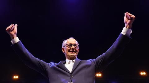 Merz steht lächelnd vor einem schwarzen Hintergrund und breitet die Arme mit den Daumen nach oben weit aus. 