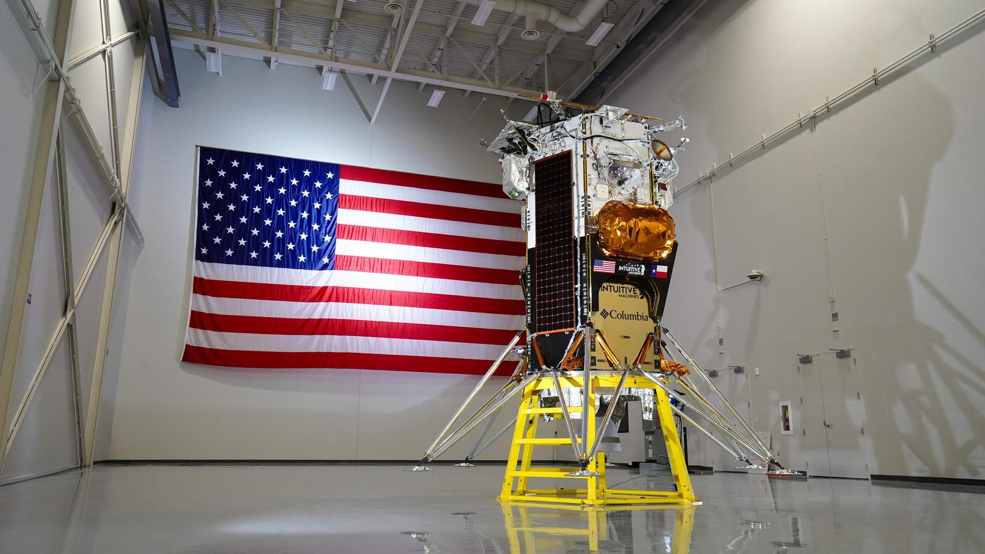 Der Lander Nova-C der Firma Intuitive Machines steht in einer Halle. An der hinteren Wand die US-Flagge.