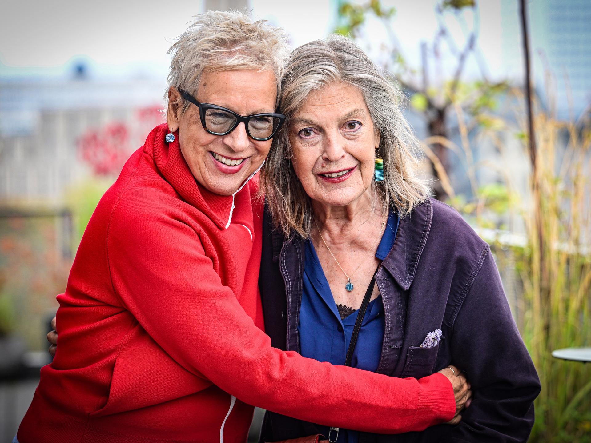 Doris Dörrie ist links im Bild auf einem Balkon, sie umarmt Angela Winkler. Die 71-jährige Dörrie trägt eine rote Jacke und hat eine Brille mit dunklen Gestell auf. Winkler hat lange Haare und trägt ein blaues Oberteil unter lilafarbener Jacke. Doris Dörrie ist links im Bild auf einem Balkon, sie umarmt Angela Winkler. Die 71-jährige Dörrie trägt eine rote Jacke und hat eine Brille mit dunklen Gestell auf. Winkler hat lange Haare und trägt ein blaues Oberteil unter lilafarbener Jacke.