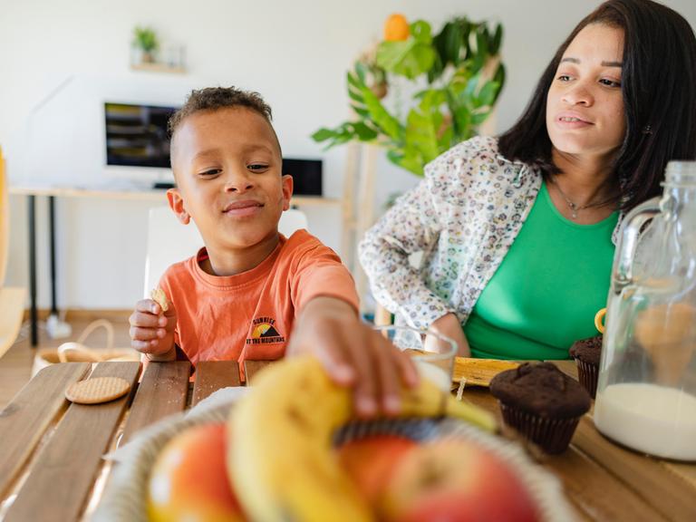 Ein Junge sitzt mit seiner Mutter am Frühstückstisch und greift nach einer Banane.