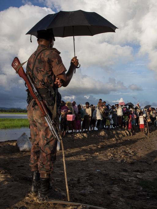 Ein Wächter aus Bangladesch mit geschultertem Gewehr steht unter einem Sonnenschirm während Tausende Rohingya-Flüchtlinge aus Myanmar an einem schlammigen Reisfeld entlanggehen.
