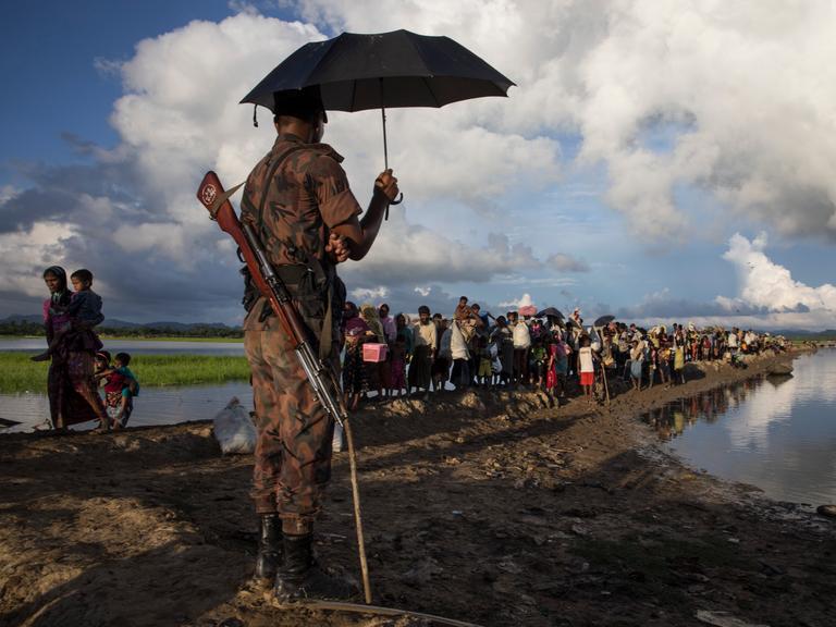 Ein Wächter aus Bangladesch mit geschultertem Gewehr steht unter einem Sonnenschirm während Tausende Rohingya-Flüchtlinge aus Myanmar an einem schlammigen Reisfeld entlanggehen.
