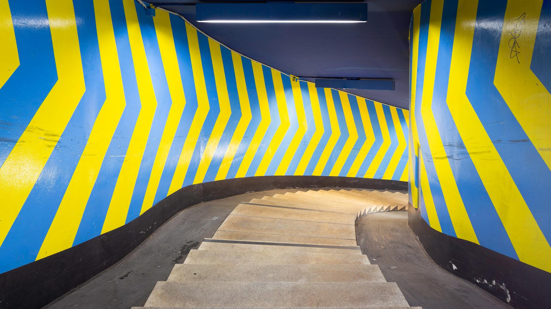 Das Bild zeigt eine enge Wendeltreppe, deren Wände mit großflächigen Pfeilen in blau und gelb bemalt sind.