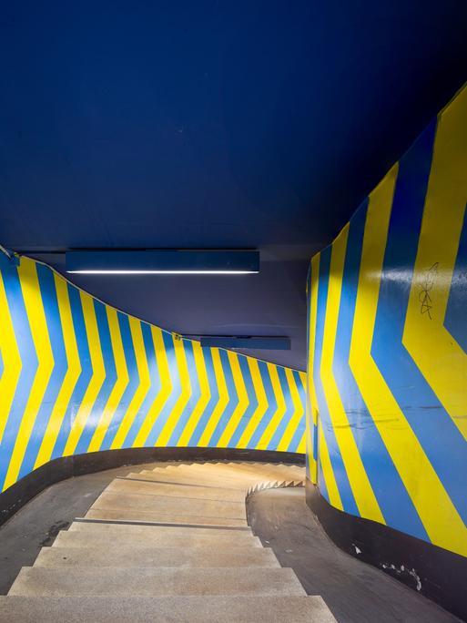 Das Bild zeigt eine enge Wendeltreppe, deren Wände mit großflächigen Pfeilen in blau und gelb bemalt sind.