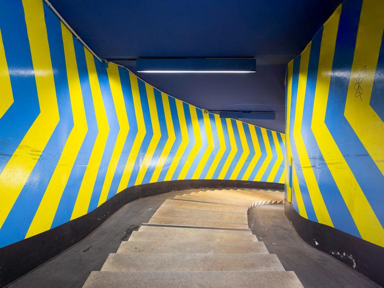 Das Bild zeigt eine enge Wendeltreppe, deren Wände mit großflächigen Pfeilen in blau und gelb bemalt sind.