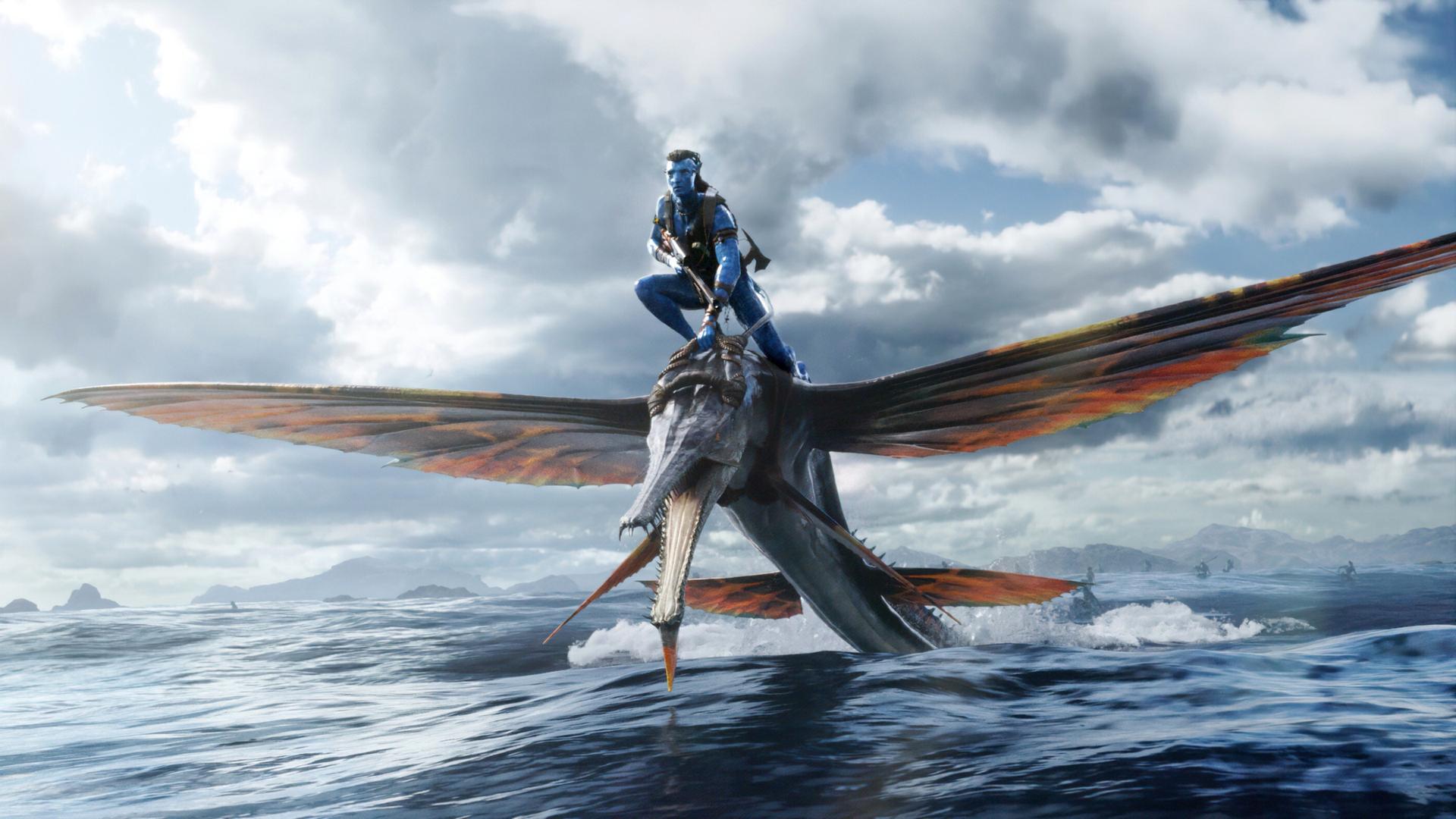 Ein Filmfoto aus James Camerons Fantasyfilm "Avatar: The Way of Water". Ein Angehöriger des Na'vi-Volkes reitet auf einem flugsauierähnlichen großen Tier über das Wasser. Die Na'vi haben blaue Haut und lange Haare.