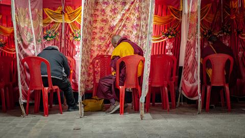 Zwischen aus Stoff aufgespannten Wahlkabinen sitzen zwei Mönche in traditioneller Kleidung, daneben eine Person in normaler Straßenkleidung. Sie sind von hinten zu sehen.