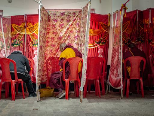 Zwischen aus Stoff aufgespannten Wahlkabinen sitzen zwei Mönche in traditioneller Kleidung, daneben eine Person in normaler Straßenkleidung. Sie sind von hinten zu sehen.