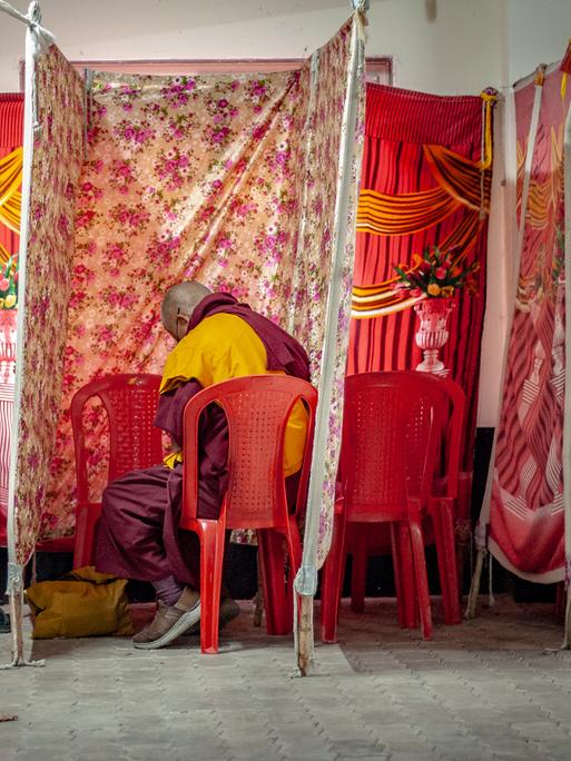 Zwischen aus Stoff aufgespannten Wahlkabinen sitzen zwei Mönche in traditioneller Kleidung, daneben eine Person in normaler Straßenkleidung. Sie sind von hinten zu sehen.