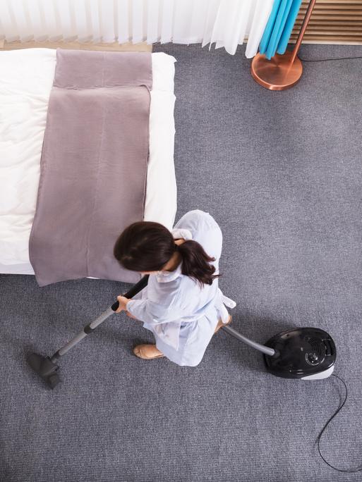 Eine Person saugt ein Hotelzimmer. Die Aufnahme ist von oben aus der Vogelperspektive gemacht.