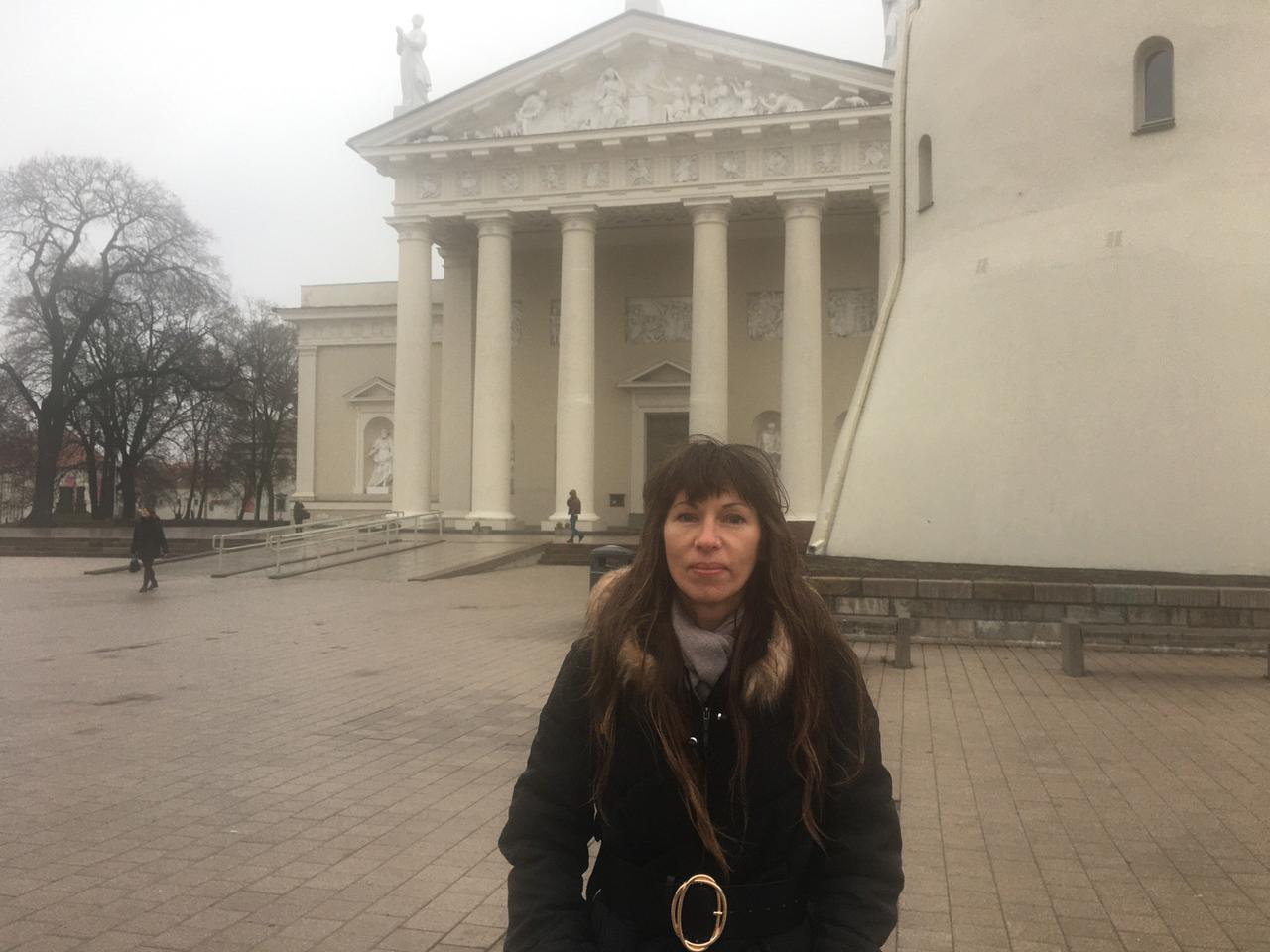 Eine Frau im Wintermantel steht auf einem Platz, im Hintergrund ein weißes Gebäude mit Säulen: Elena Kotenochkina