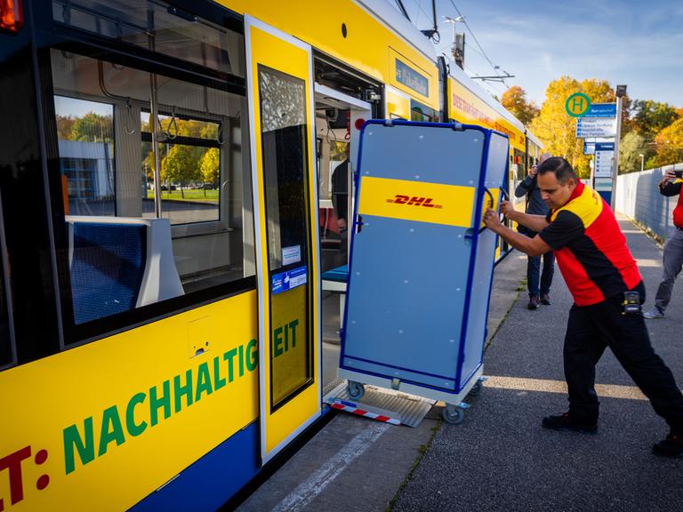 Ein Mitarbeiter der Deutschen Post schiebt einen Transportwagen für Pakete in eine Straßenbahn.