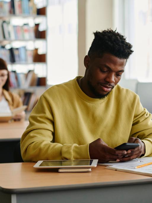 Ein junger Mann an einem Bibliothekstisch hält ein Handy in der Hand, neben ihm liegen ein Schreibblock, ein Tablet und ein Buch.