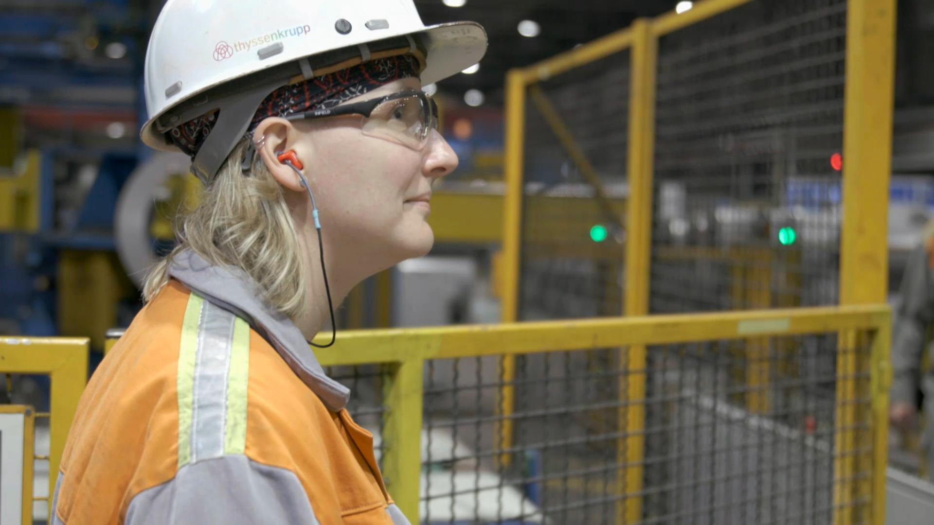 Ann Catherine Meyer steht mit Schutzhelm und Arbeitsjacke in einer Produktionshalle von Thyssenkrupp und blickt nach rechts Ann Catherine Meyer steht mit Schutzhelm und Arbeitsjacke in einer Produktionshalle von Thyssenkrupp und blickt nach rechts
